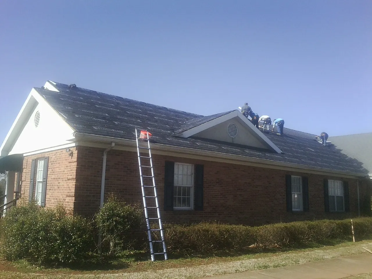 Crew tearing shingles off a brick ranch roof with a ladder set — Metal Roof Repair work by our crew in Cerrillos