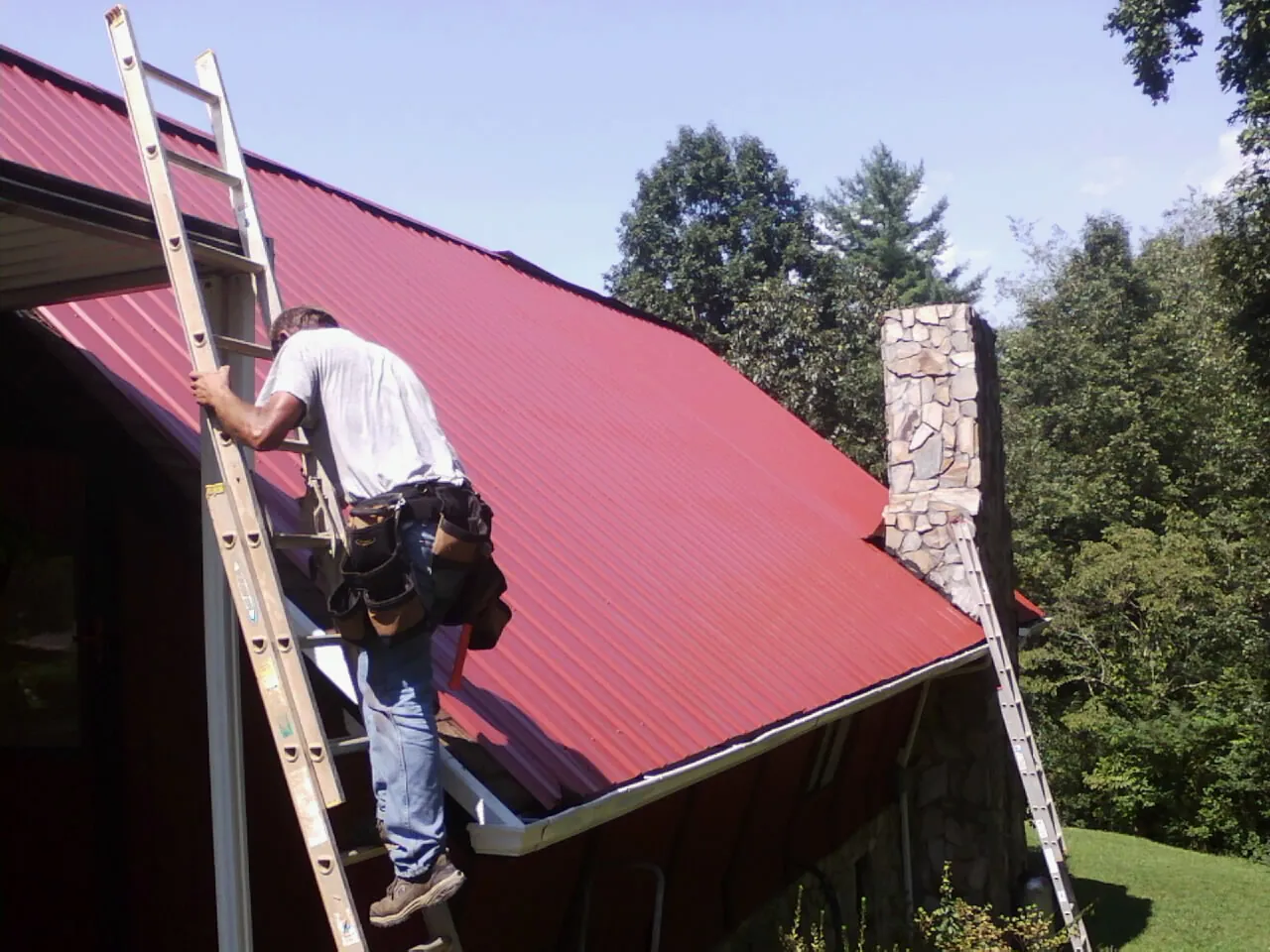 Roofer on a ladder beside a red metal roof and stone chimney — Soffit & Fascia Repair by Roofing Brothers in Cerrillos